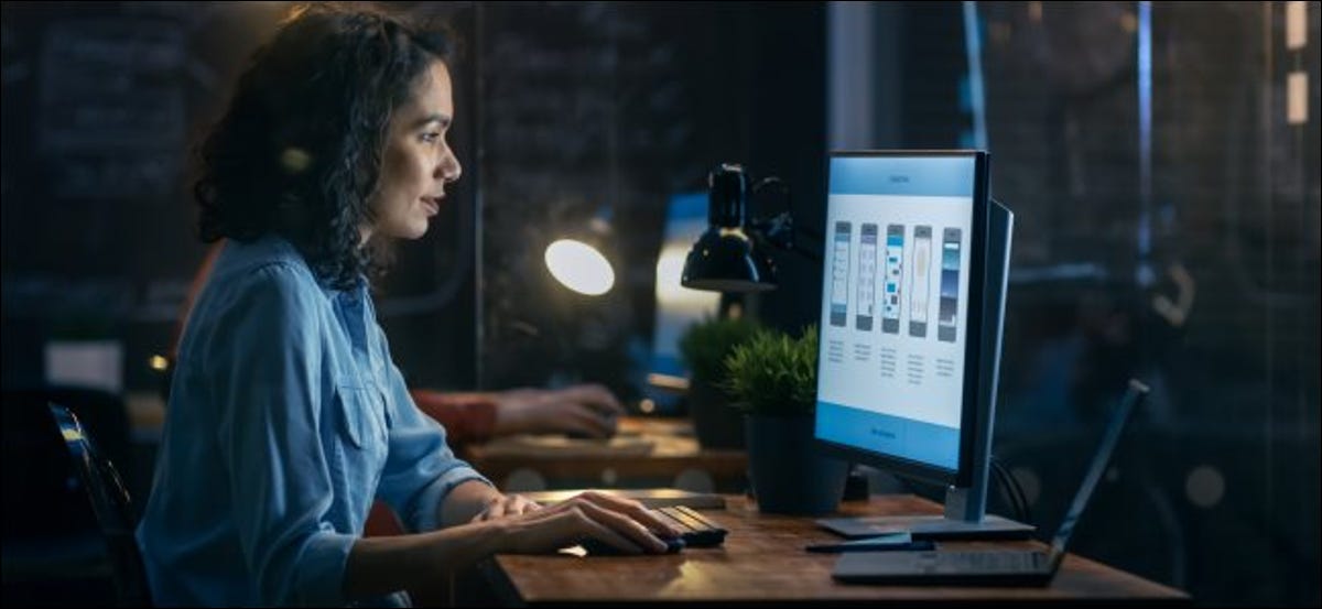 Una mujer que trabaja en una computadora de escritorio, con una computadora portátil abierta al lado. 