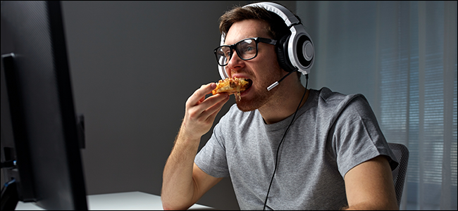 Un hombre sentado frente a una computadora, con auriculares y comiendo una pizza.