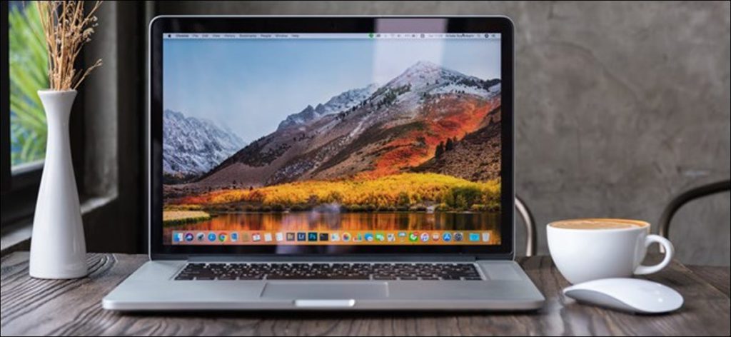 Un MacBook Pro abierto sobre una mesa junto a una taza y un jarrón de flores.