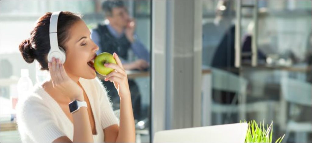 Una mujer que usa audífonos mientras usa un Apple Watch y come una manzana.