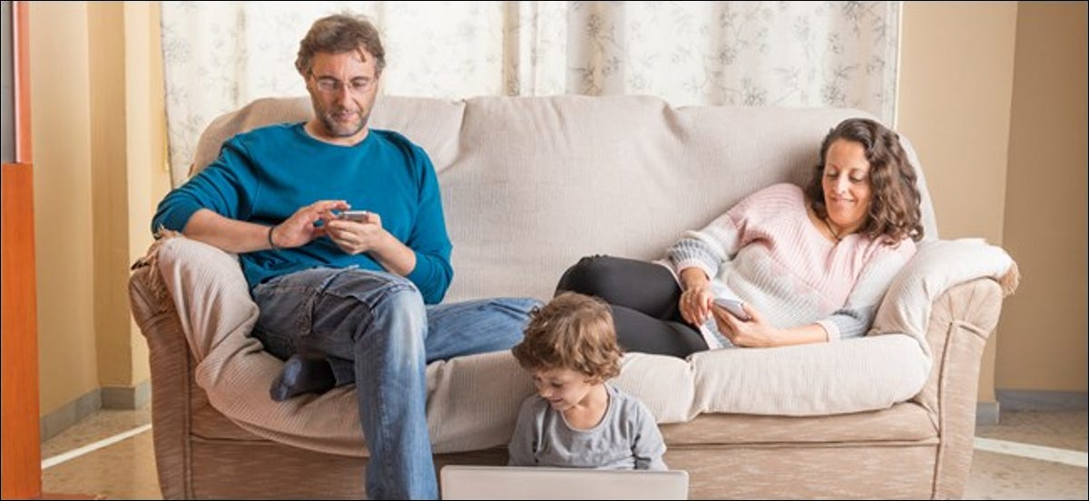 Un hombre y una mujer en un sofá miran sus teléfonos, mientras que un niño pequeño se sienta en el suelo junto a ellos con una computadora portátil abierta.